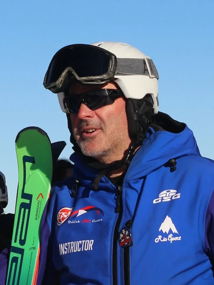 A man wearing a helmet, sunglasses, and a blue ski jacket outdoors, holding a green ski, against a clear blue sky.