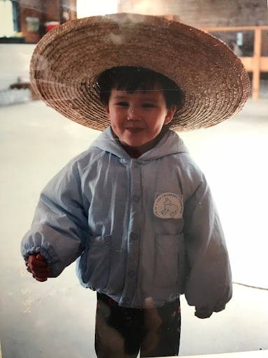A young boy wearing a large woven straw hat and a light blue jacket, smiling indoors.