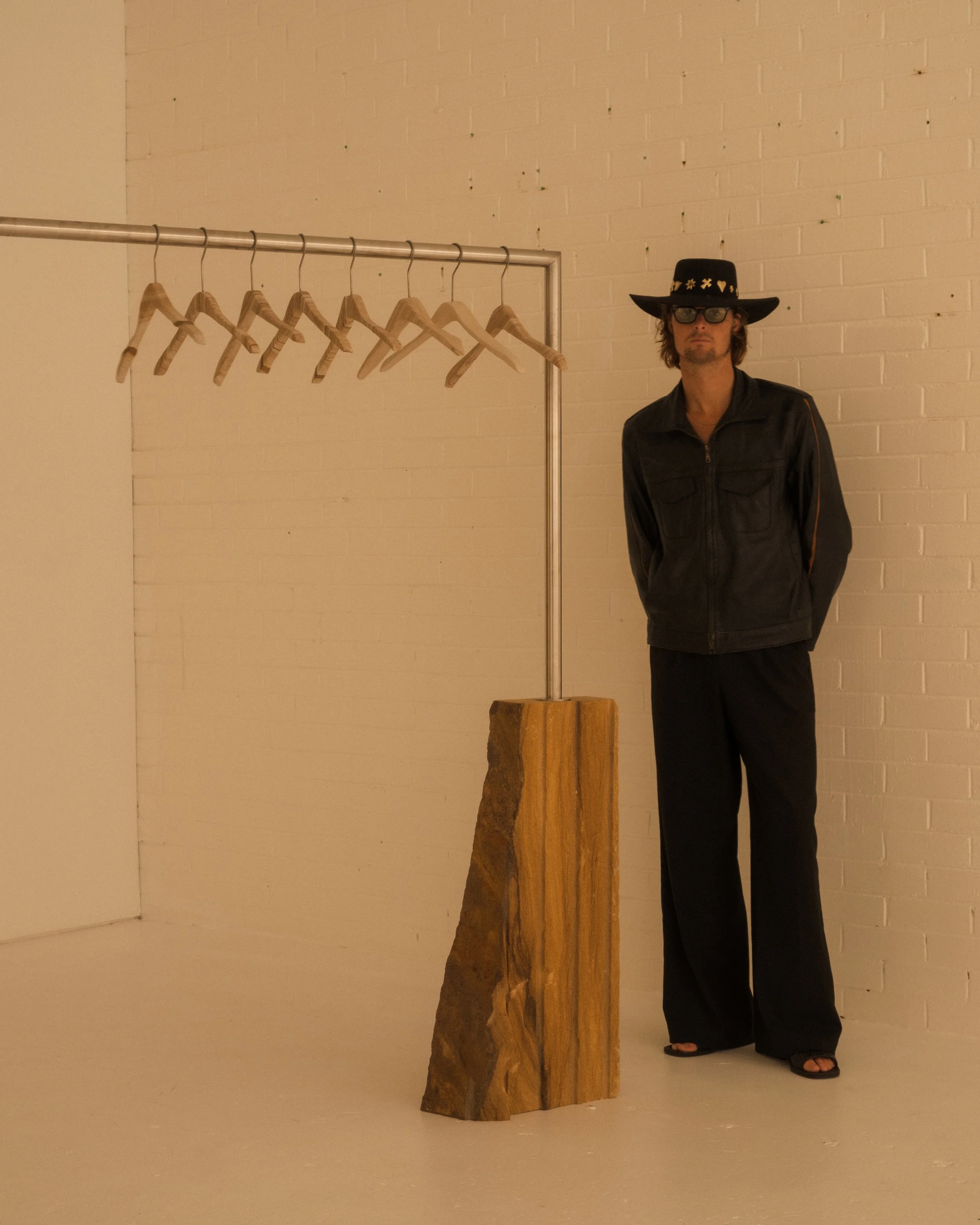 Person wearing a black hat, sunglasses, and a black leather jacket standing next to a clothing rack with wooden hangers in a white room.