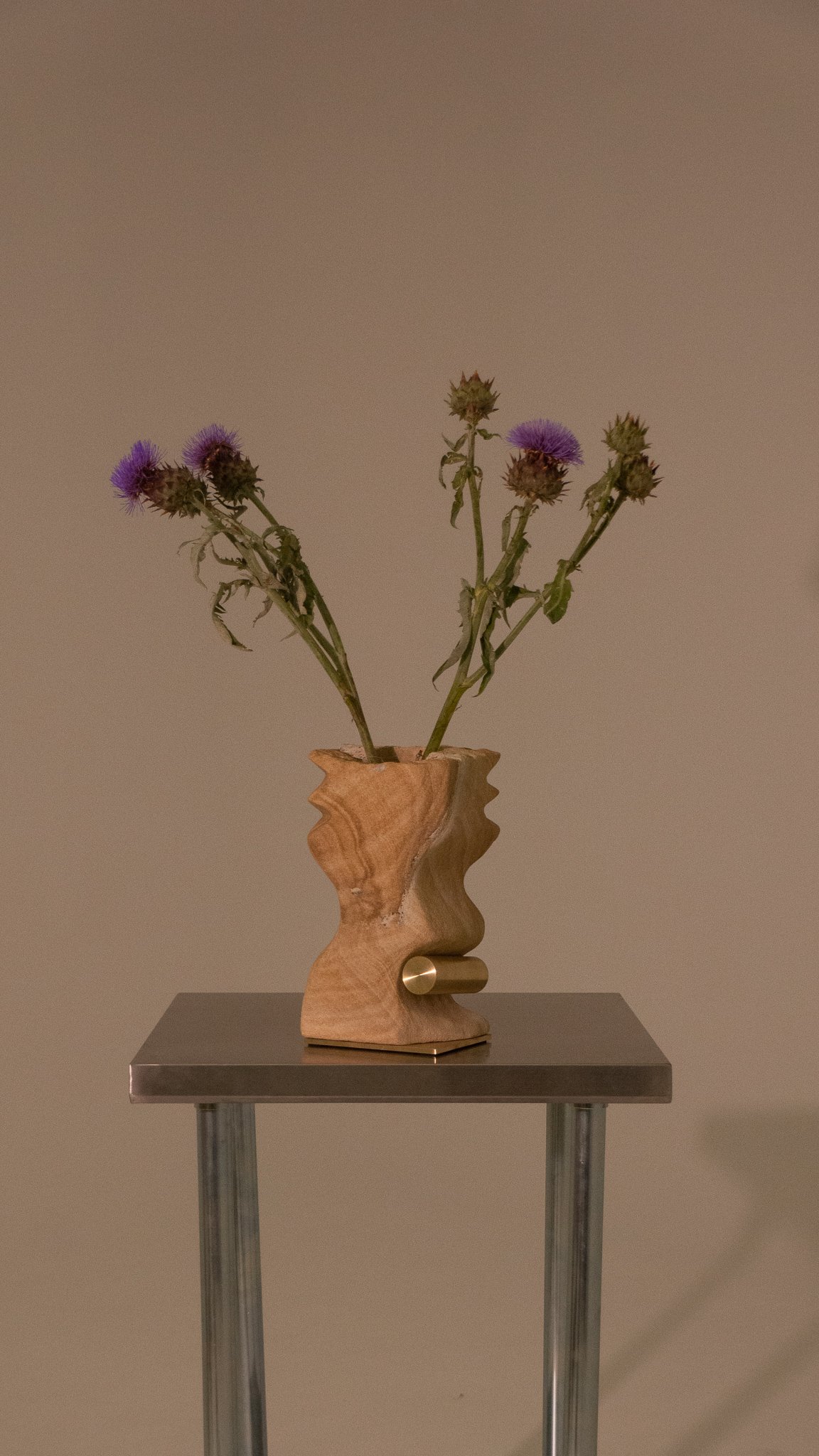 A wooden vase with an irregular shape holding purple wildflowers, placed on a metal stand against a plain wall.