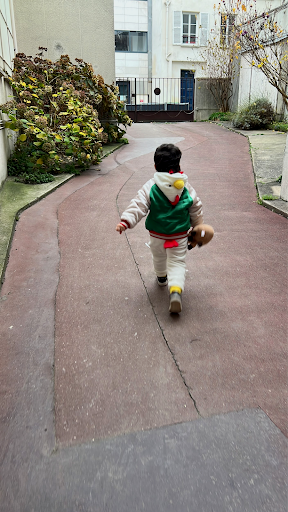 Child running down a curved pathway holding a football, wearing a green and white jacket with a hood, in an outdoor courtyard with plants and buildings.