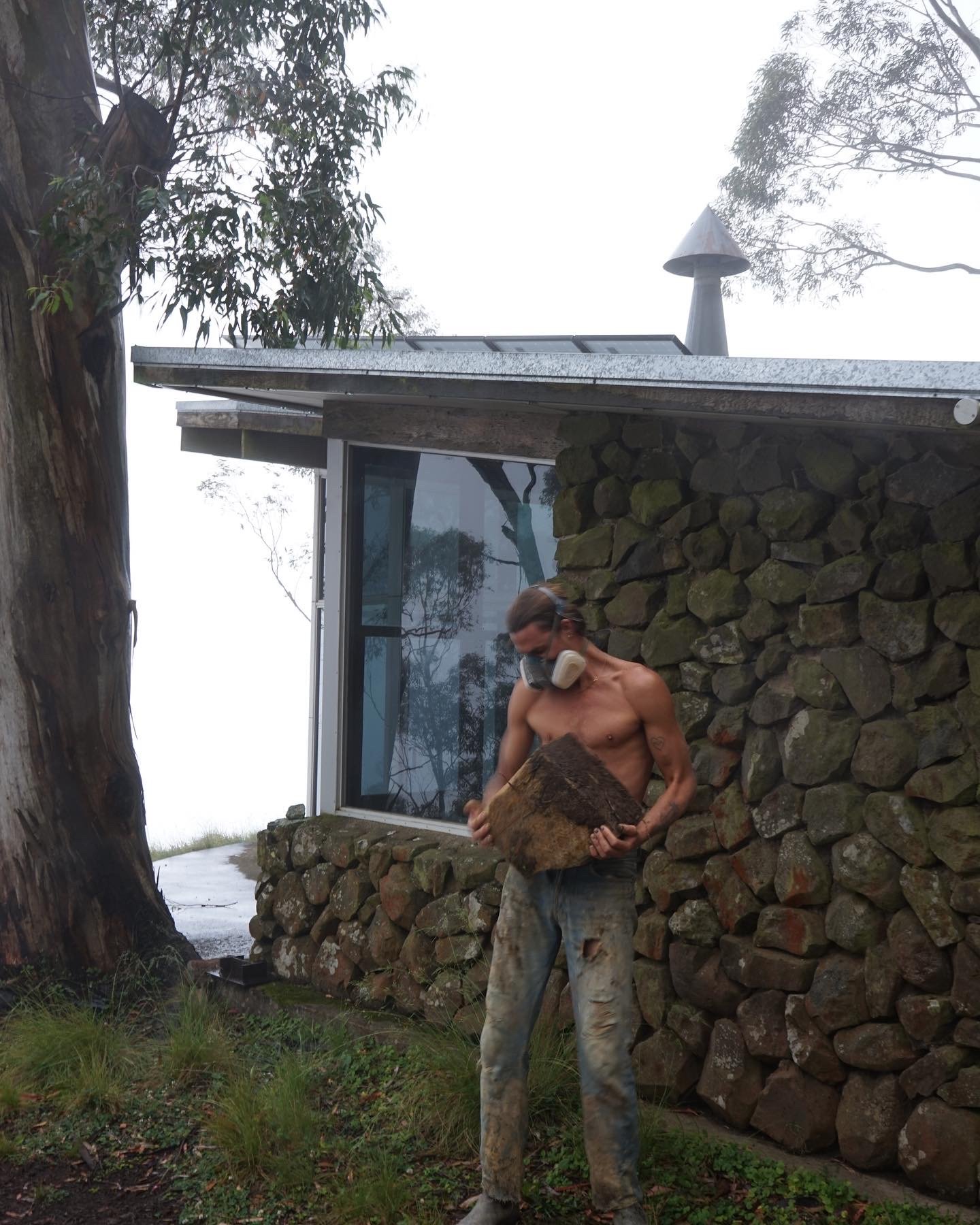 A shirtless man wearing glasses and a respirator mask is holding a large piece of wood or bark outside a stone house with a large window. There are trees and fog in the background.