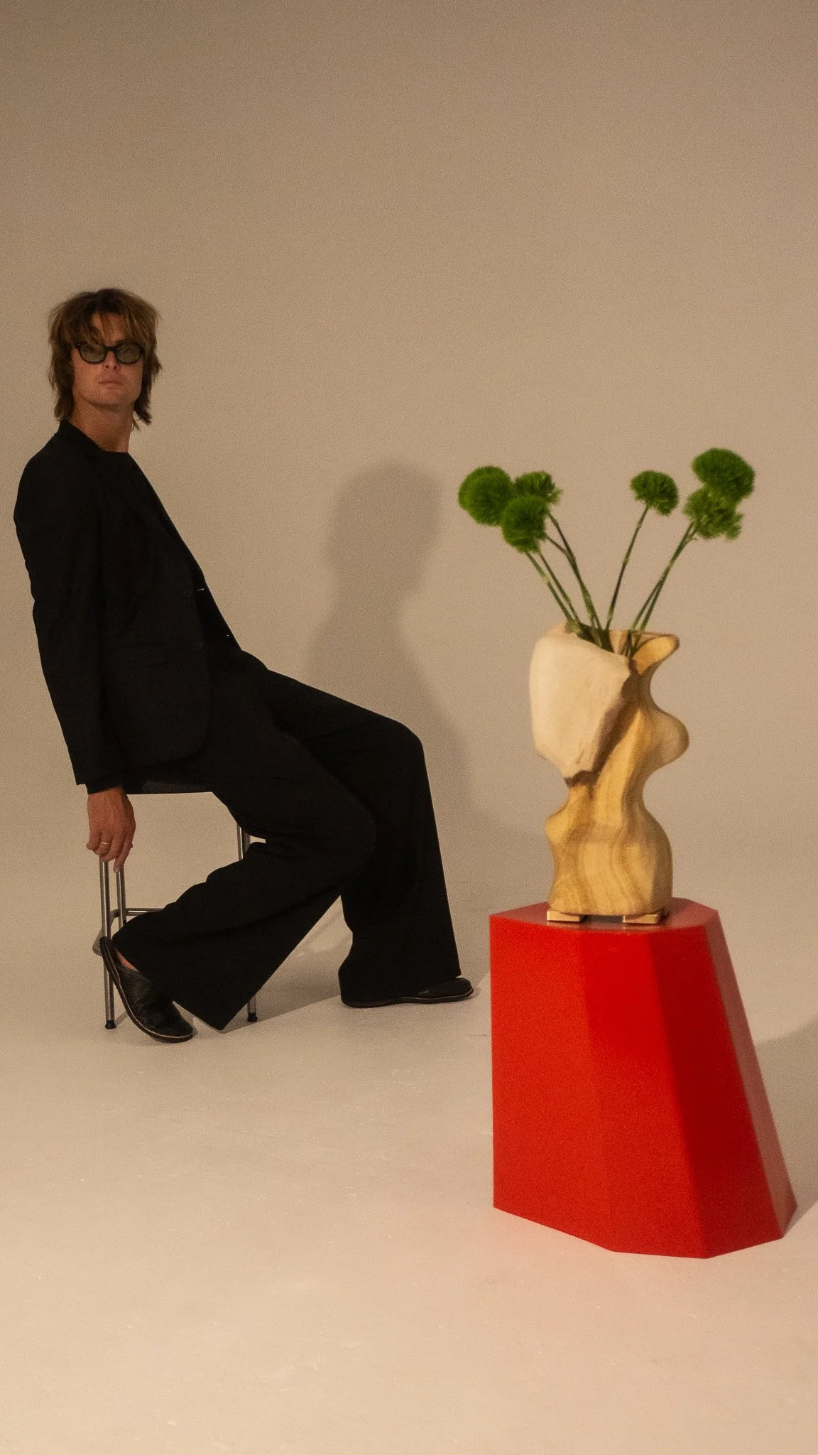 A person sitting on a stool with a black suit and sunglasses next to a red pedestal with a wooden sculpture of a vase holding green spherical flowers against a plain background.