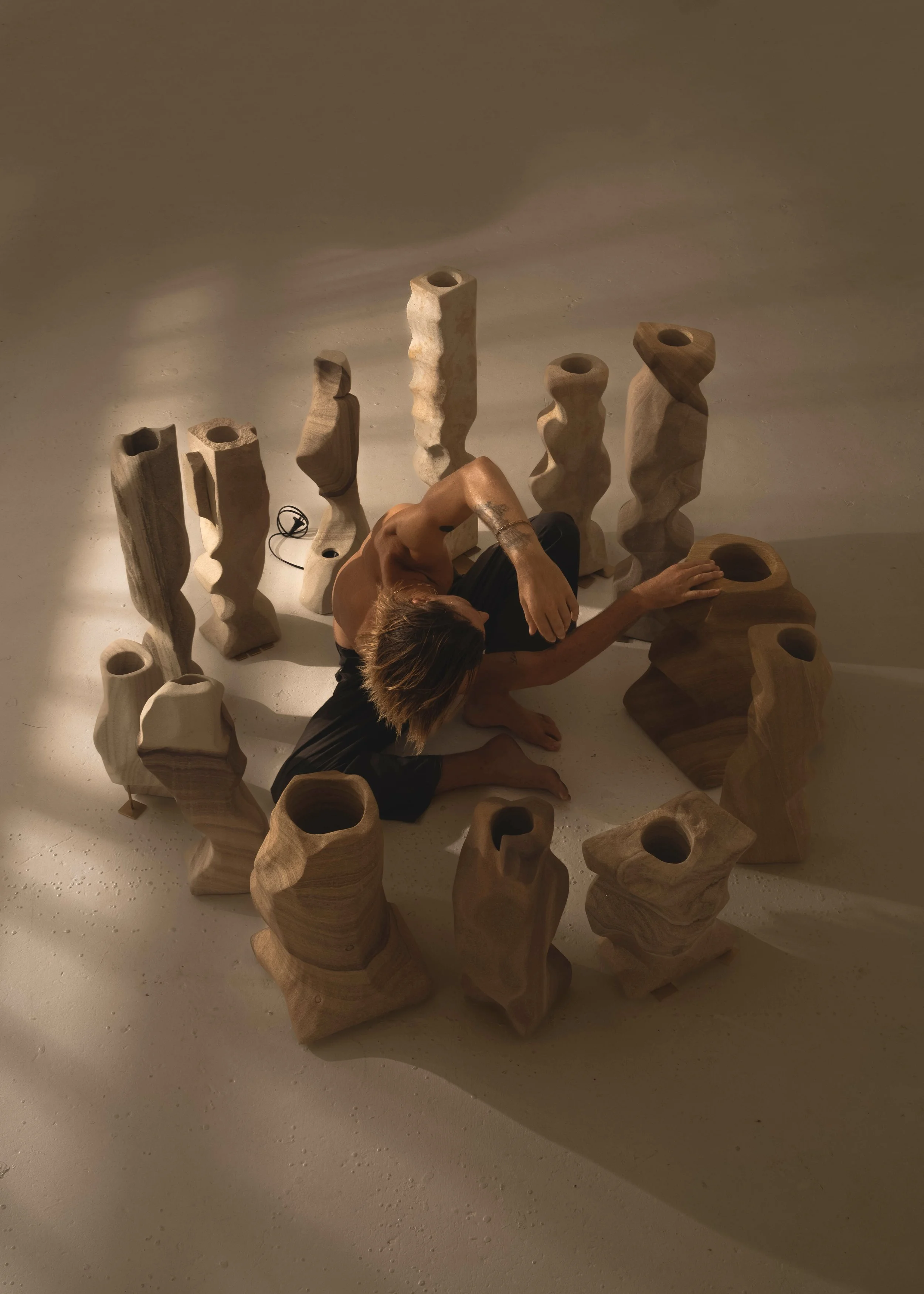 A person with brown hair is kneeling on the ground surrounded by multiple abstract wooden sculptures with holes and wavy designs, reaching out to one of the sculptures.