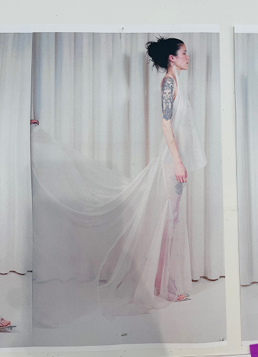 A woman with dark hair styled in an updo, standing sideways in front of cream-colored curtains, wearing a sheer white dress with a long train and visible tattoos on her arm.