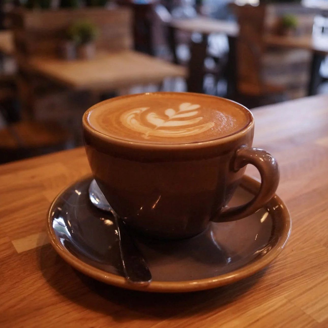 A brown ceramic cup filled with a latte art coffee drink on a matching saucer with a spoon, placed on a wooden table in a cozy cafe with blurred background of empty chairs and tables.