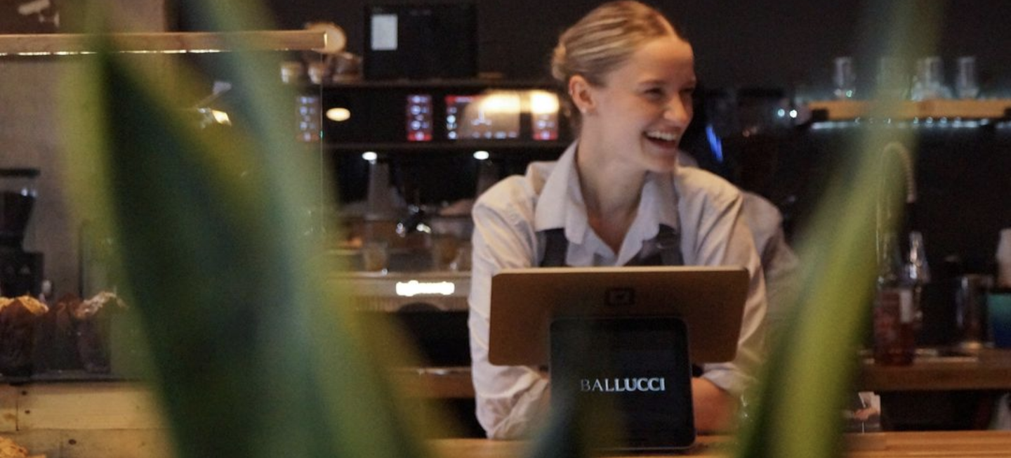A woman with blonde hair tied back, wearing a white shirt, smiling and working at a restaurant or cafe counter with a laptop labeled "Ballucci."