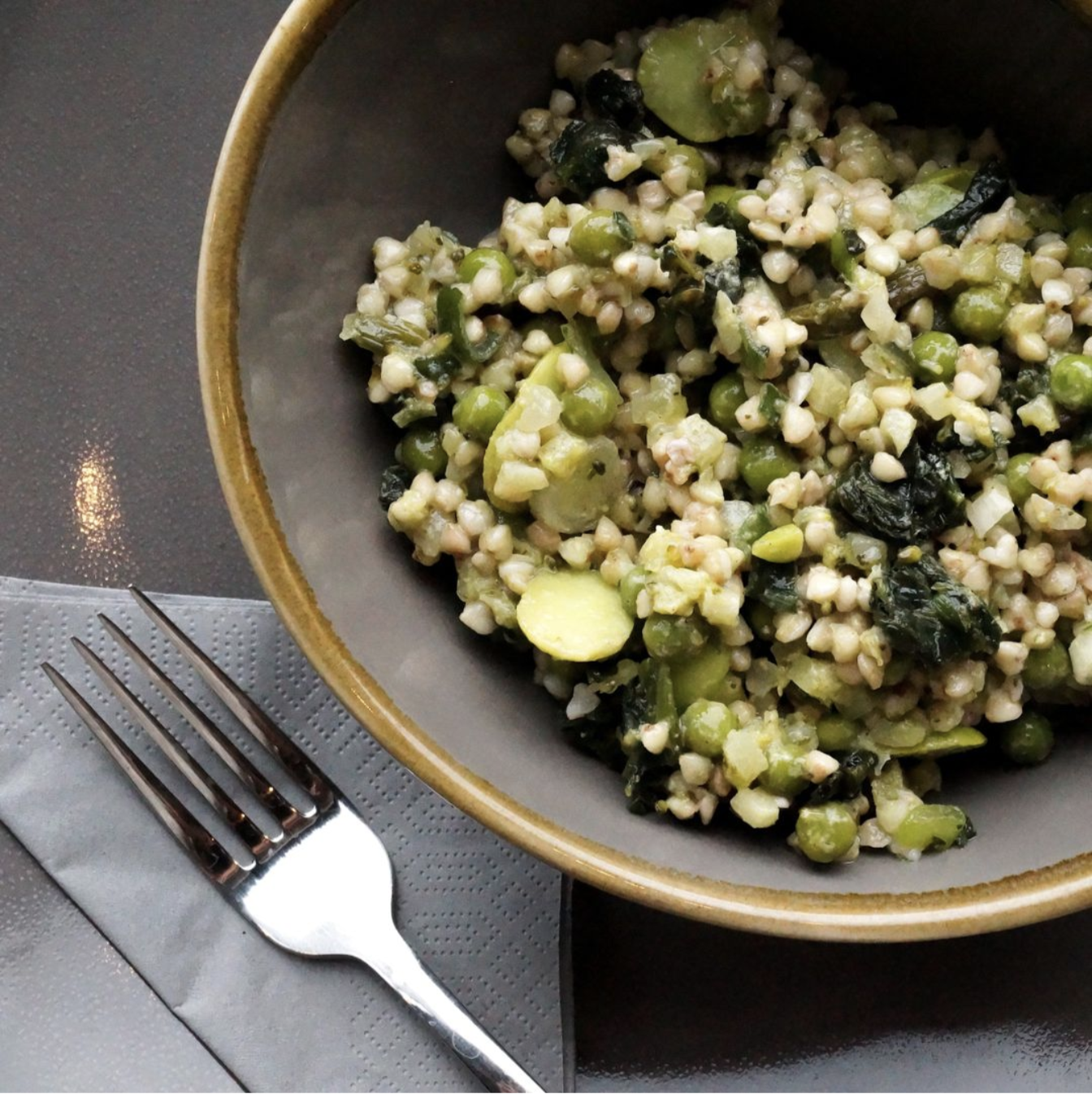 A bowl of cooked couscous mixed with chopped vegetables and leafy greens, placed on a gray table with a fork on a cloth napkin.