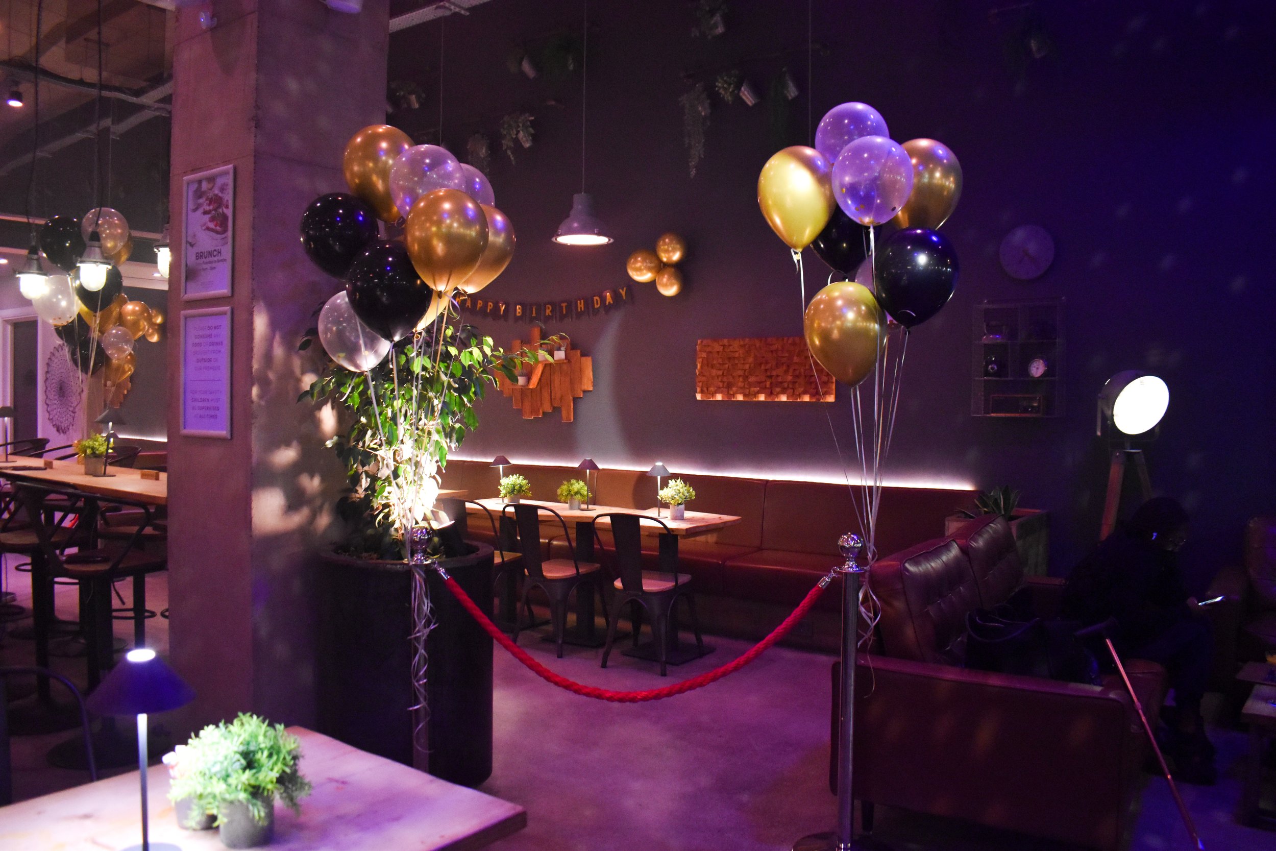 Indoor setting decorated for a celebration with black, gold, and clear balloons, potted plants, and a 'Happy Birthday' banner, with dining tables and chairs.