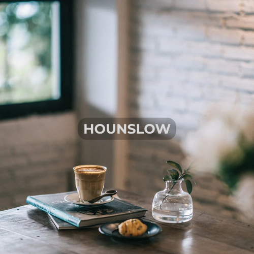 A cup of coffee, a plate with a croissant, a glass vase with a green plant, and a spoon on a table near a window and brick wall.