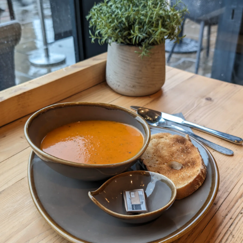 A bowl of orange-colored soup, a slice of toasted bread on a dark plate, a small pitcher of cream or milk, and silverware, all placed on a light wooden table near a window with a potted plant in the background.