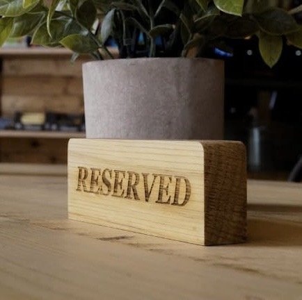 A wooden block with the word 'RESERVED' engraved on it, placed on a wooden table in front of a potted plant.