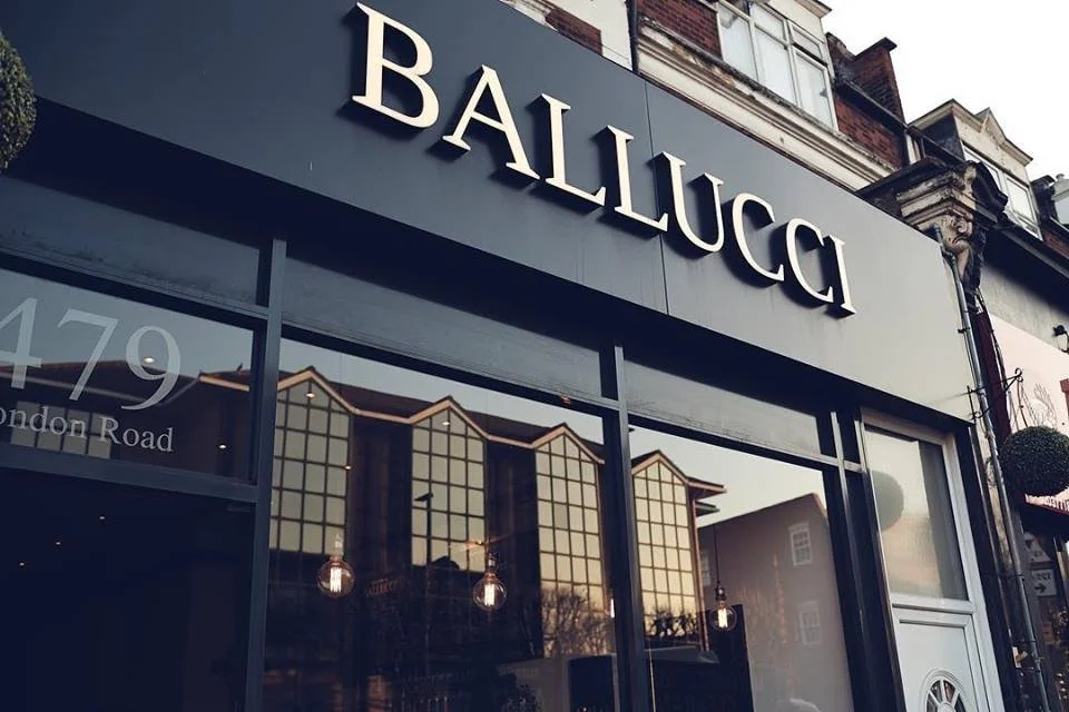 Storefront sign reading 'BALIUCCI' with large windows and reflection of buildings and trees