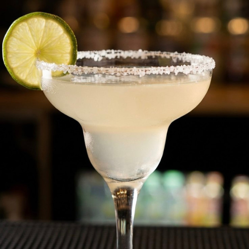 A margarita glass filled with a pale yellow cocktail, garnished with a lime wedge and rimmed with salt.