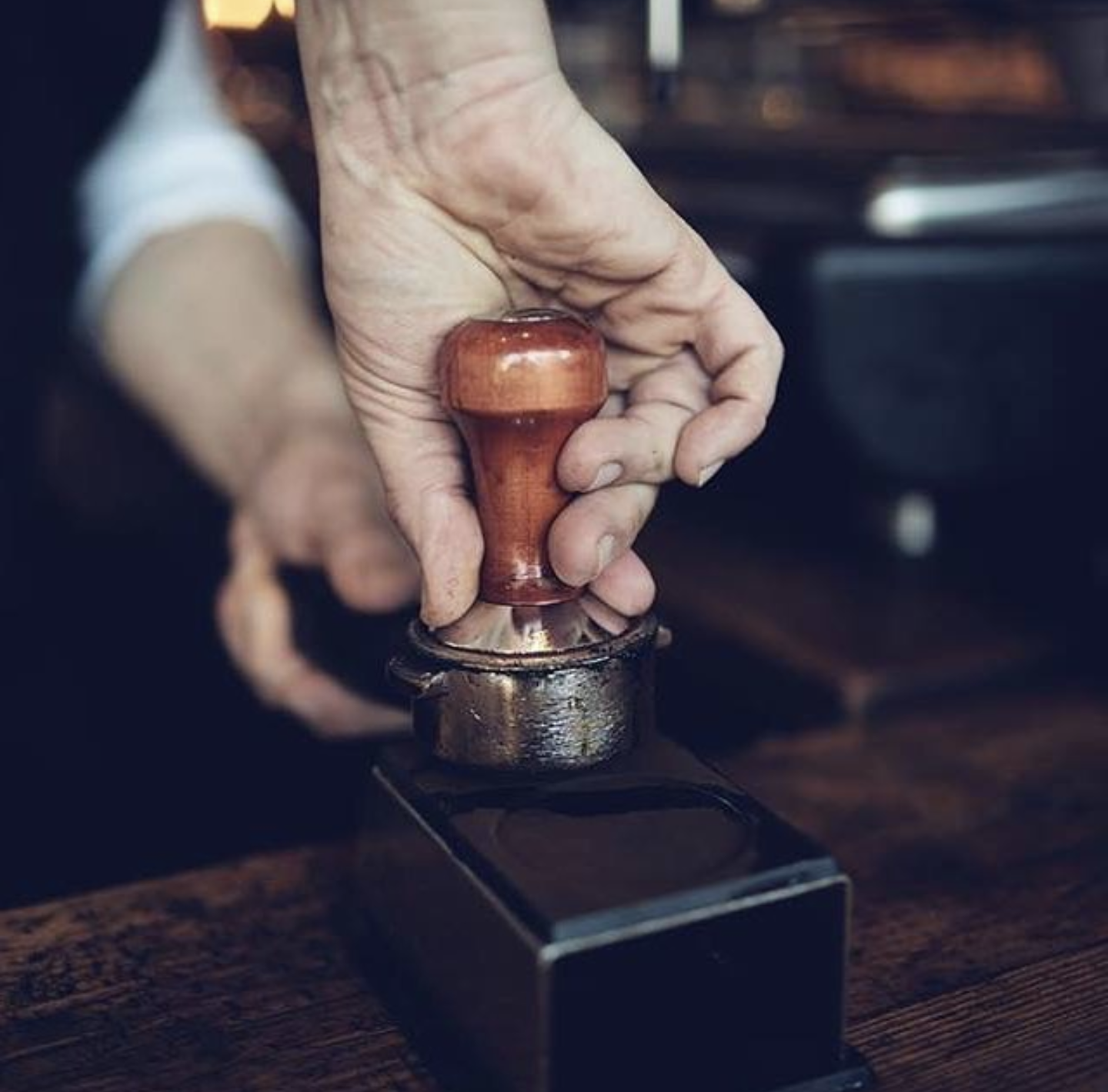 Close-up of a hand with worn skin pressing down on a wooden tamper with a metal base, likely used for coffee or espresso, on a dark wooden surface.