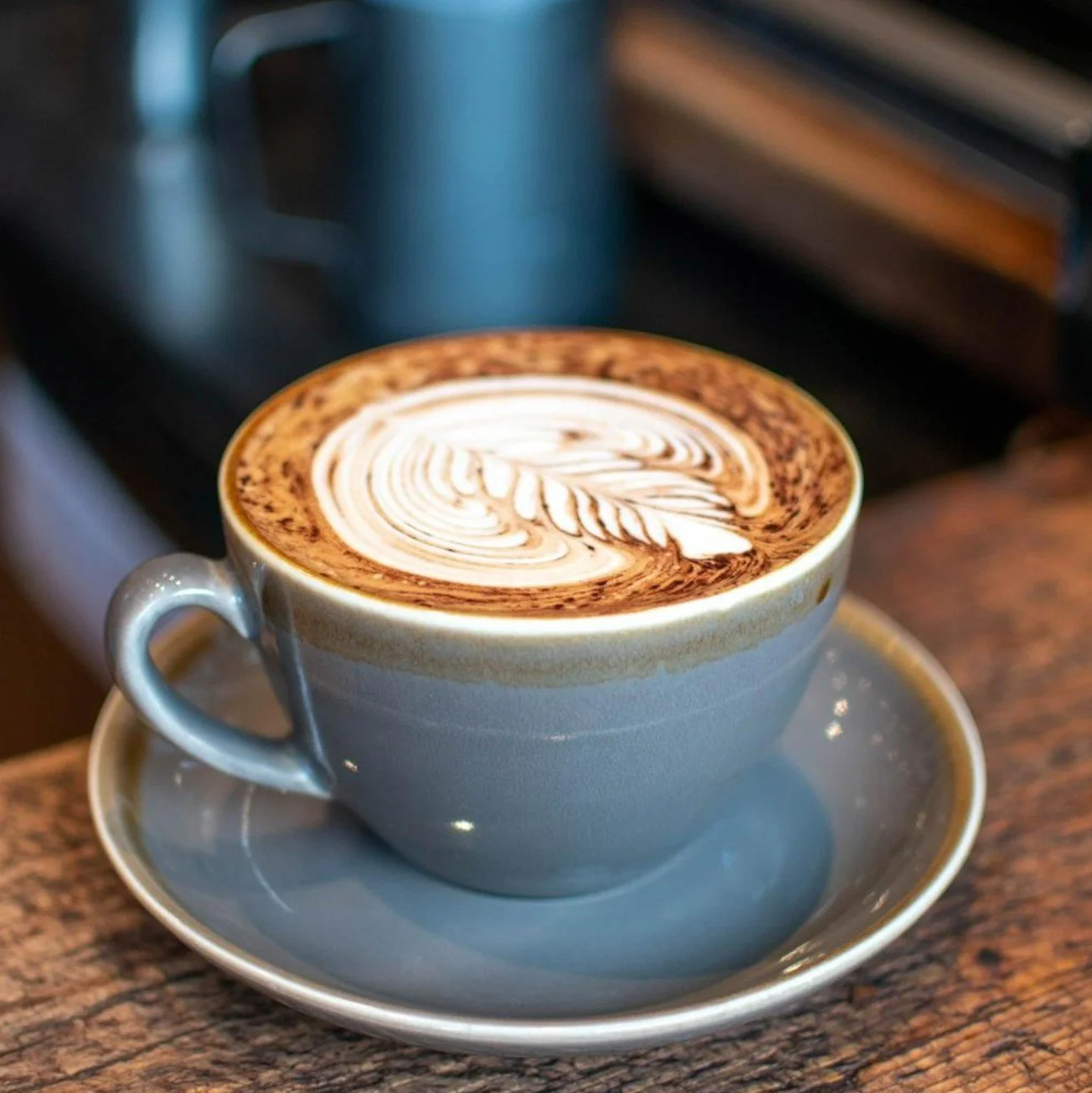 A blue ceramic cup with a matching saucer containing a cappuccino with latte art on top, placed on a wooden surface.