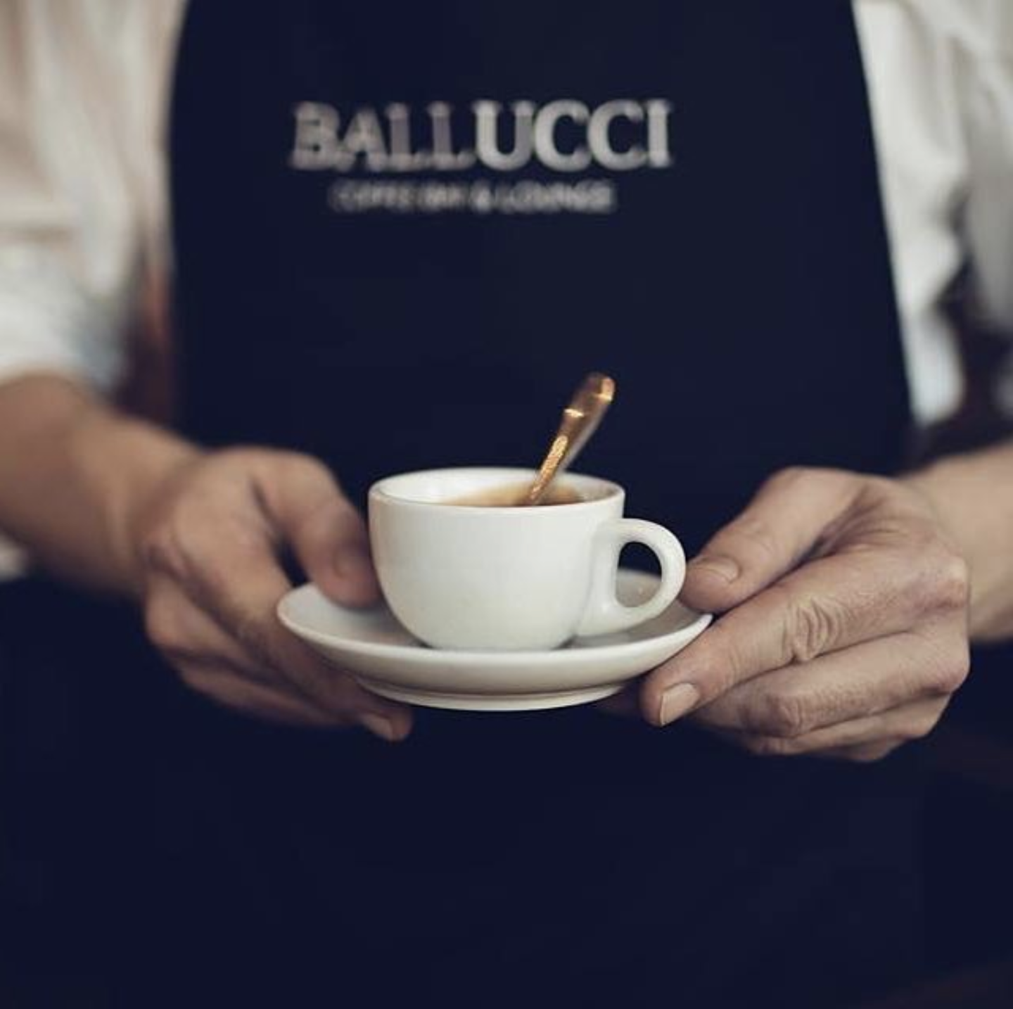 Person holding a small white coffee cup with a saucer, a spoon inside, wearing a black apron with blurred text.