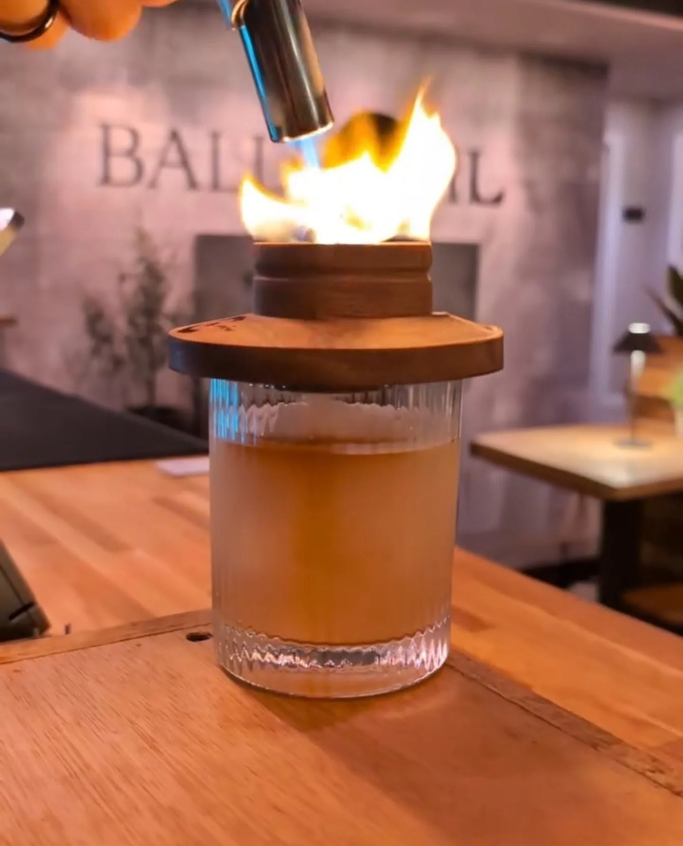 A flame torches on a glass container with a wooden lid, placed on a wooden table in a restaurant or café setting.