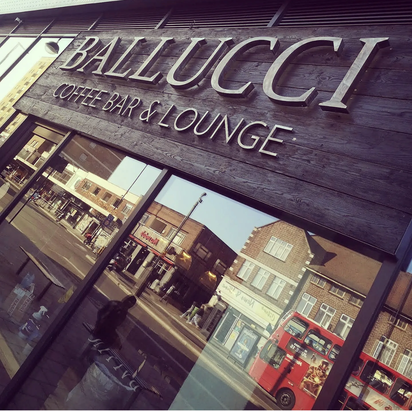 View of a coffee shop and lounge named 'Ballucci' with a wooden sign, reflected on glass windows, with street scene and buildings in the background, including a red double-decker bus.