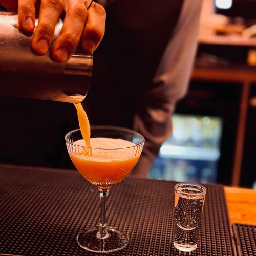 A bartender pouring a bright orange cocktail into a coupe glass on a bar counter with a shot glass nearby.