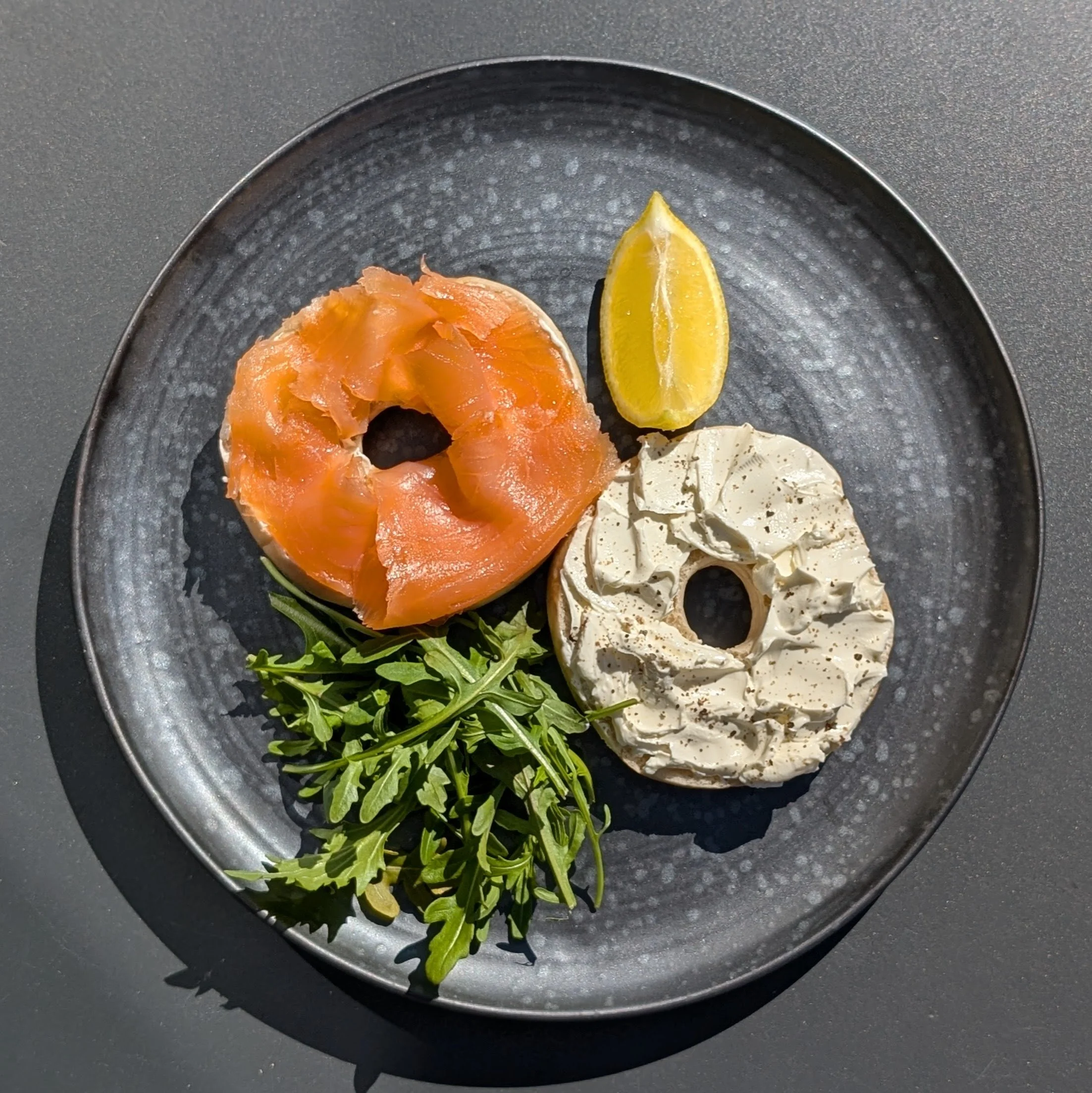 A black round plate with smoked salmon, a lemon wedge, some cream cheese with herbs, and a small bunch of arugula salad.