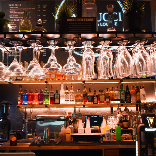 Bar counter with hanging wine glasses, colorful liquor bottles, and bar equipment in a dimly lit lounge.
