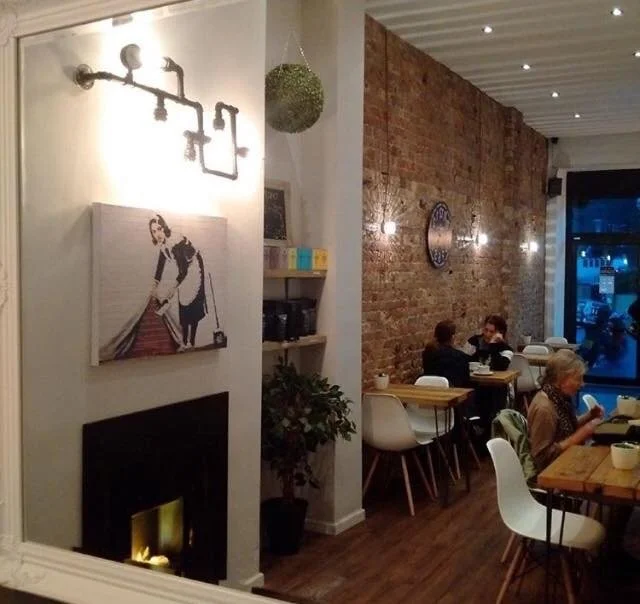 Cozy cafe interior with exposed brick wall, modern seating, wall clock, and artwork of a child and woman by a fireplace. Several customers are seated, enjoying their meals or conversations.
