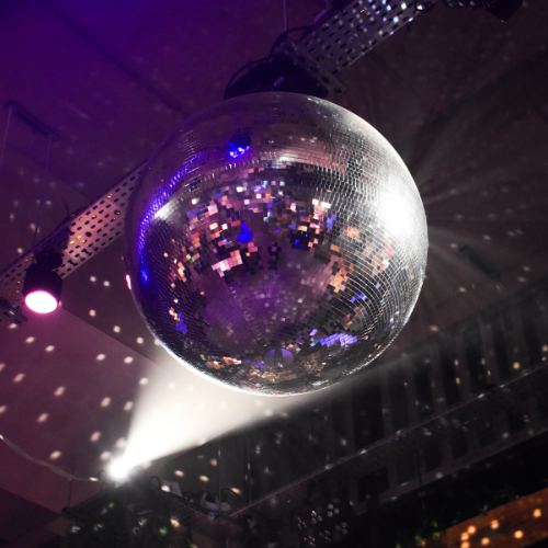 A large disco ball hanging from the ceiling, reflecting colorful lights, with a spotlight shining on it in a darkened room.