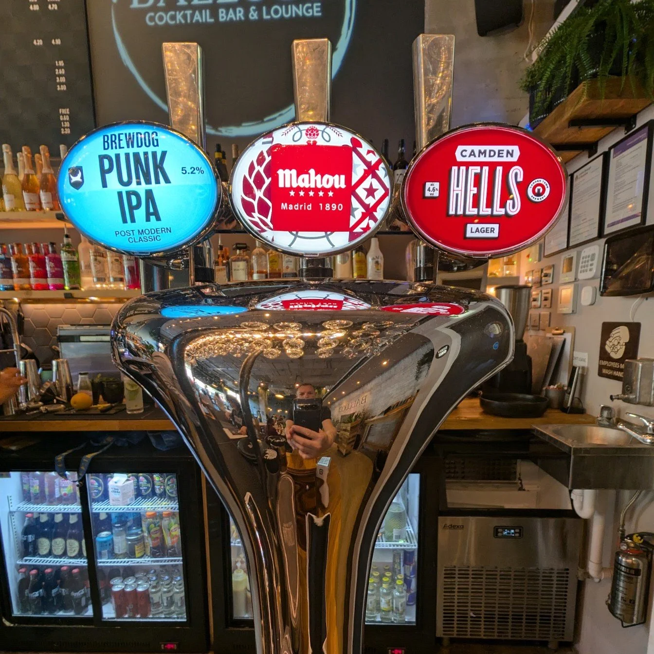 This is a photo of a bar tap with three beer options: Brewdog Punk IPA, Mahou Madrid 1890, and Camden Hells Lager. The background includes a bar counter, a fridge with beverages, liquor bottles, and a person taking a selfie reflected in the shiny sur