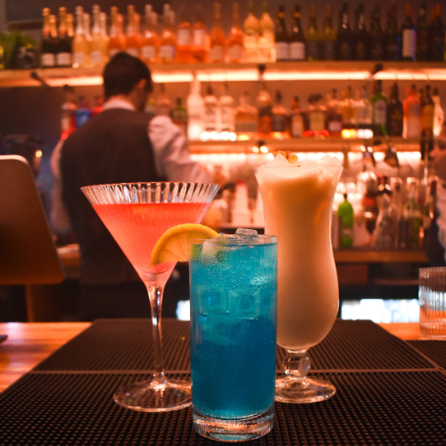 Three colorful cocktails on a bar counter with a bartender in the background and shelves of bottles