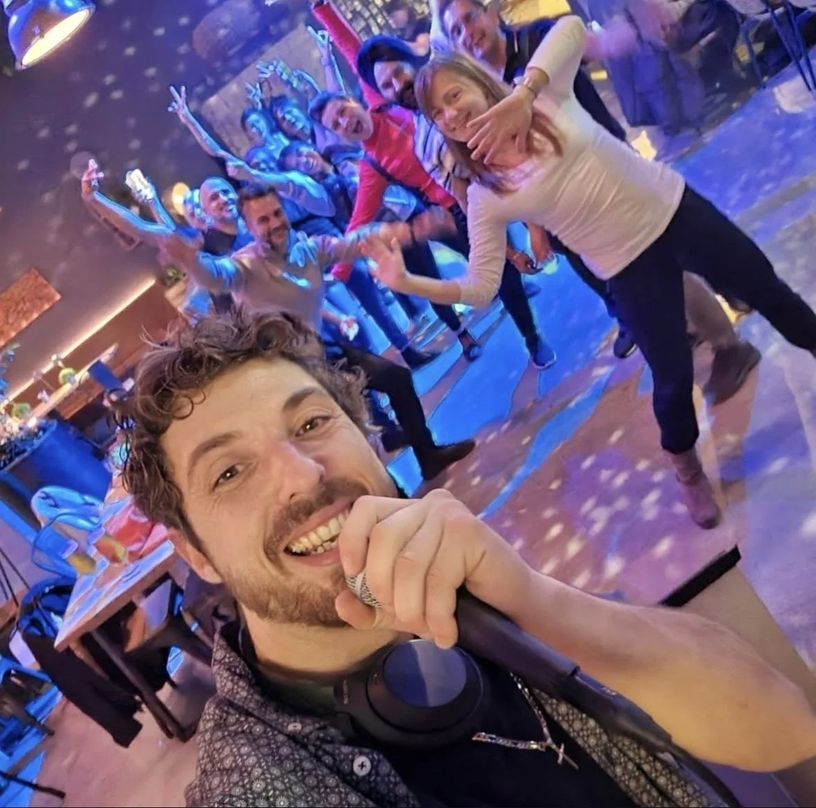 A group of people enjoying a karaoke singing session in a lively, colorful bar or club. One man is in the foreground holding a microphone, smiling, with headphones around his neck. The group behind him is laughing, singing, and celebrating, creating 