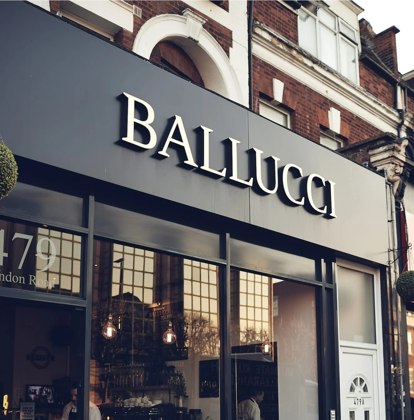 Exterior view of a storefront with a large sign that reads 'BALLUCCI' above the entrance. The store has large glass windows, reflecting buildings and trees outside. There are people inside, and the store appears to be a café or tea shop. The building above the storefront is made of brick with traditional architecture.