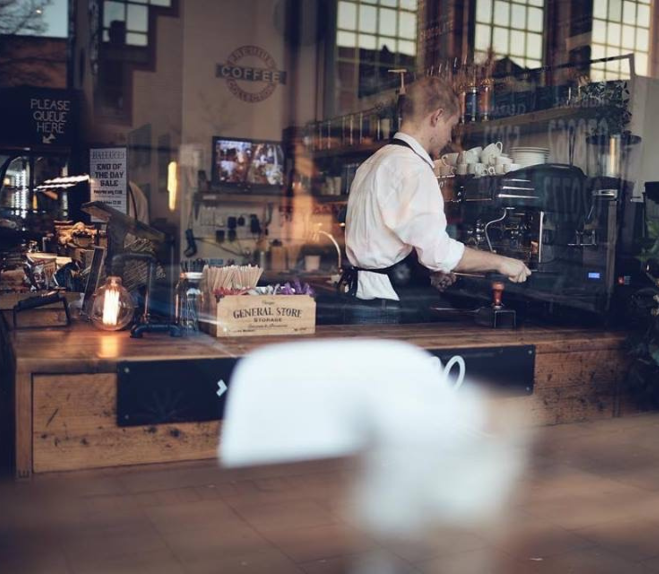 A barista in a white shirt and black apron making coffee behind a wooden counter in a coffee shop, viewed through a window.