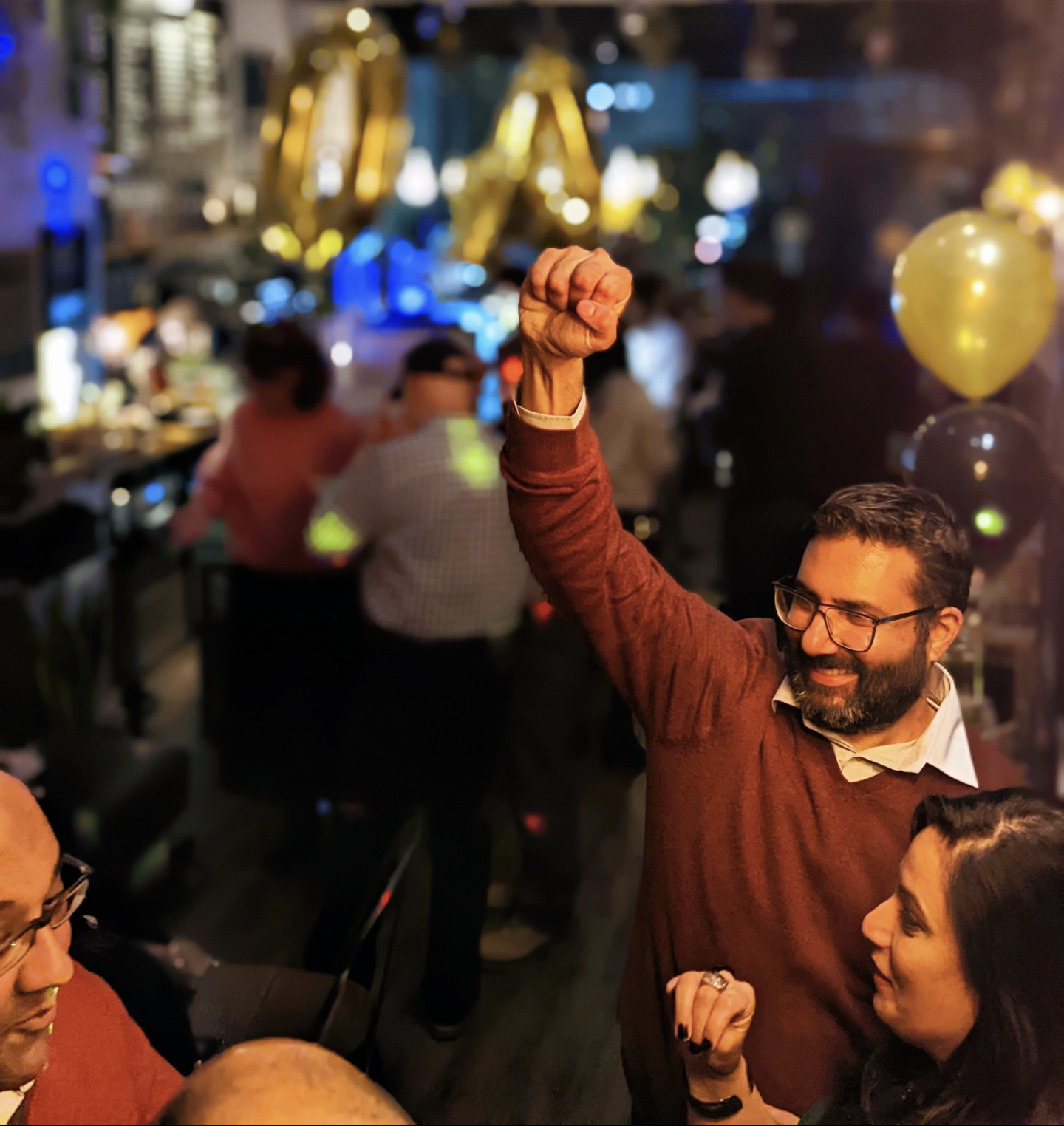 A man with glasses and a beard sitting with a woman, both smiling and holding hands, in a lively celebration with balloons and golden decorations in the background.
