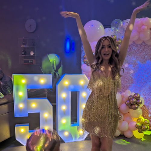 A woman in a gold, fringed dress celebrates at a 50th birthday party, standing in front of illuminated '50' sign and colorful balloons.