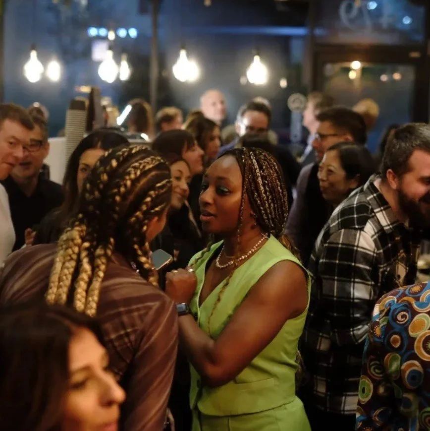 Group of people socializing at a crowded indoor event, with two women in the foreground engaged in conversation amid warm lighting.