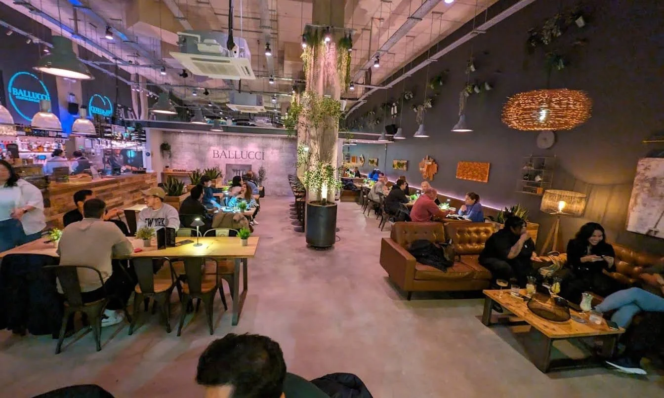 Interior of a restaurant with people dining at tables and on couches, with plants and wall decor, a central tree, and a bar area on the left side.