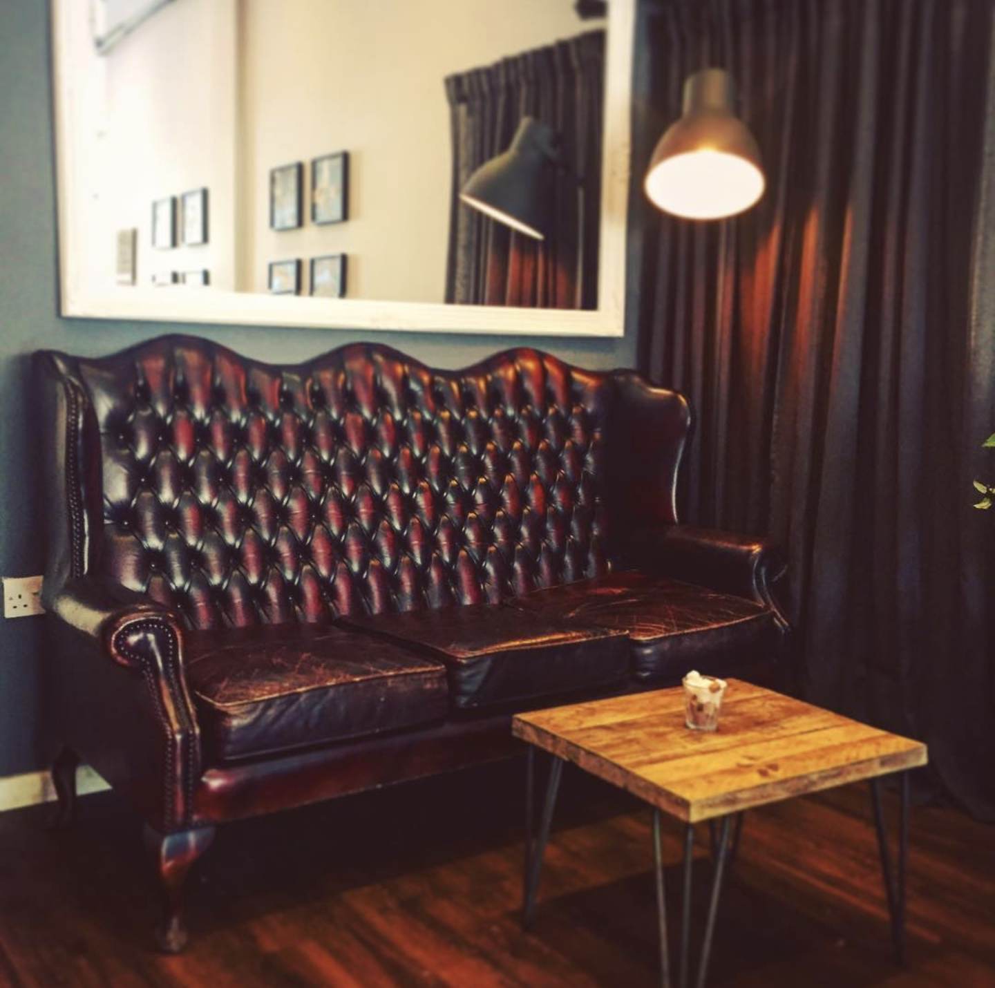 A vintage style living room corner with a dark leather tufted sofa, a small wooden coffee table with a drink on it, framed pictures on the wall, a large mirror, dark curtains, and a wall-mounted lamp.