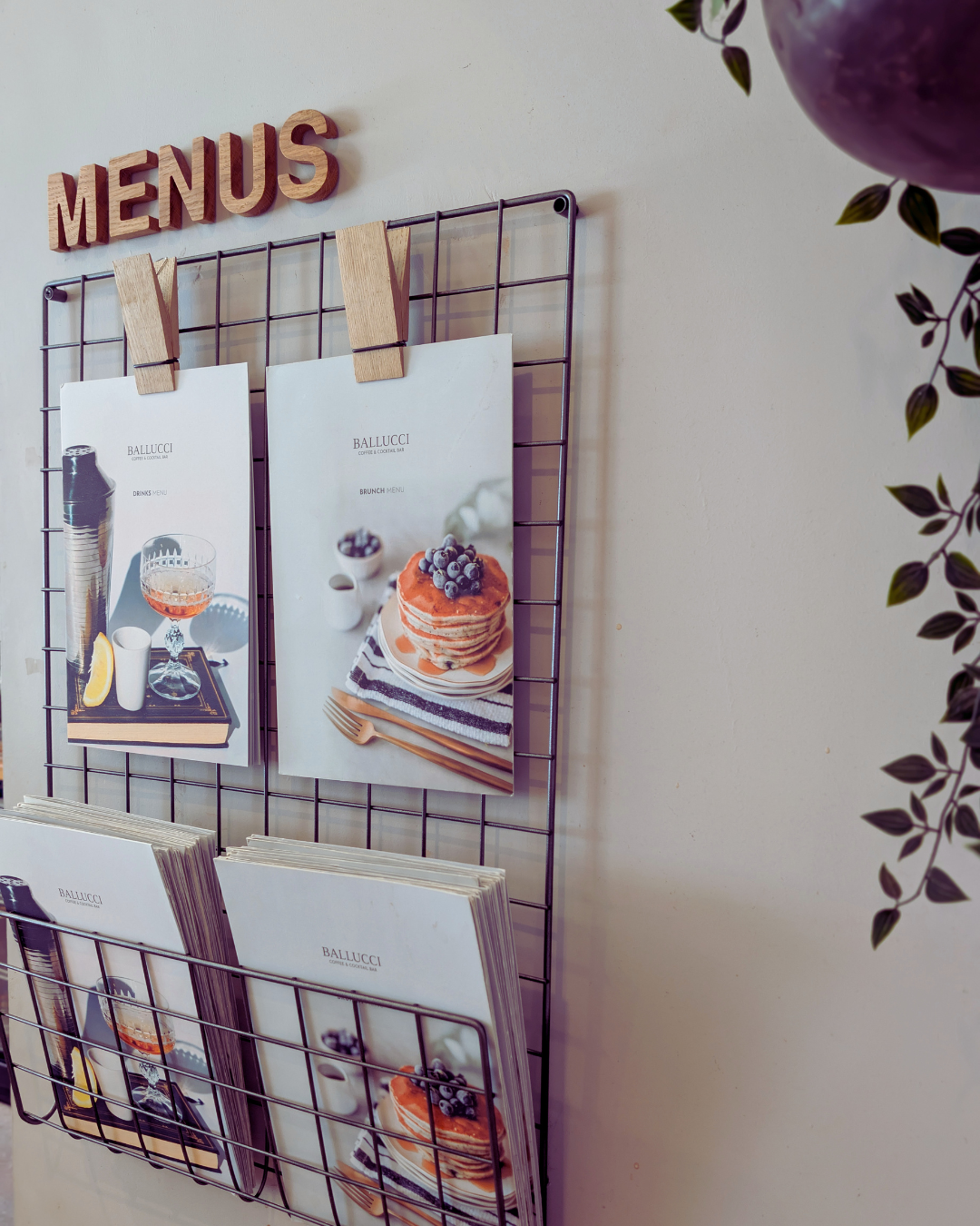 Menu display with two menus attached to a wire grid, one showing drinks with a cocktail glass and lemon, the other showing a stack of pancakes with blueberries, on a white wall.
