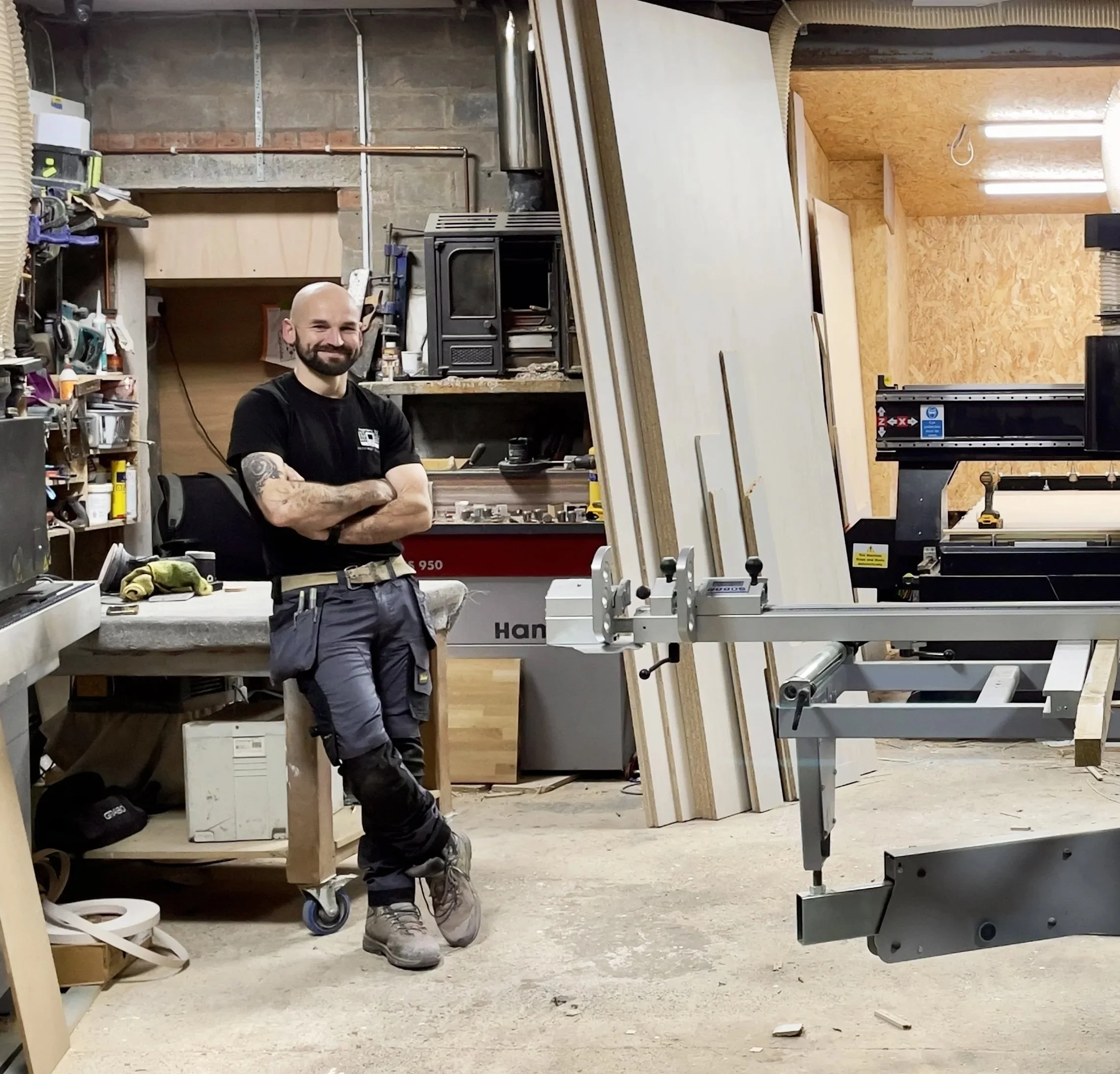 A man with a beard, tattooed arm, black t-shirt, and work pants stands confidently in a woodworking workshop, surrounded by tools, wood pieces, and equipment.
