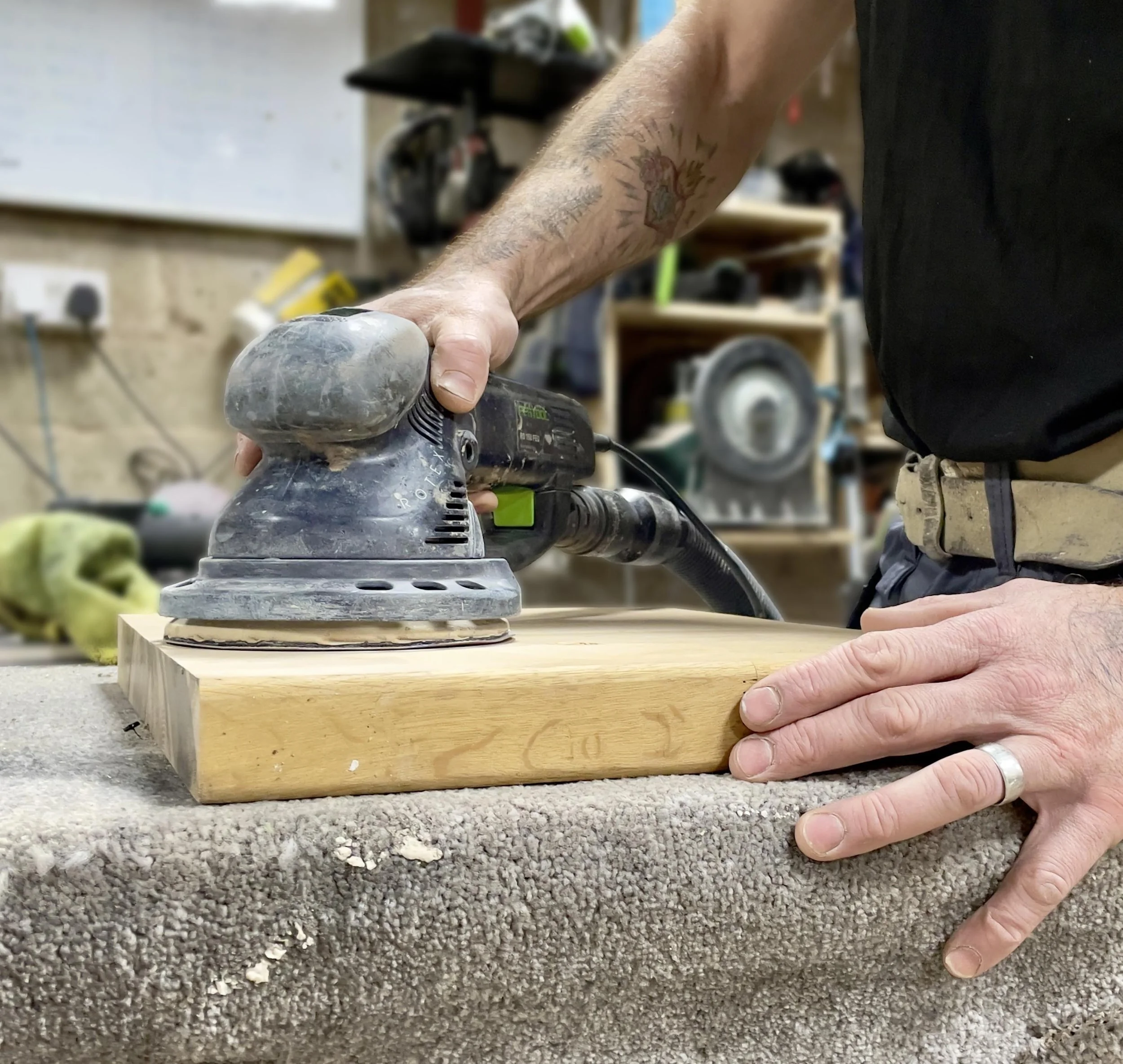 A person is sanding a piece of wood using a handheld electric sander in a workshop. The person has tattoos on their arm, and they are wearing a black shirt and beige belt. The workshop has shelves with various tools and equipment in the background.