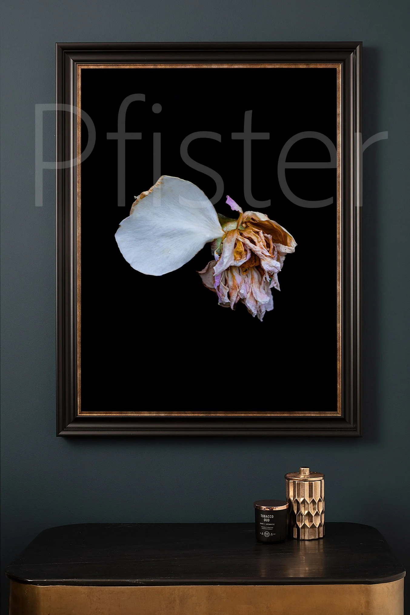 Decorative art featuring a dried white flower against a black background, framed in a black and gold frame, hanging on a gray wall above a black and gold table with a geometric candle holder and a black candle labeled 'Tobacco Oud.'