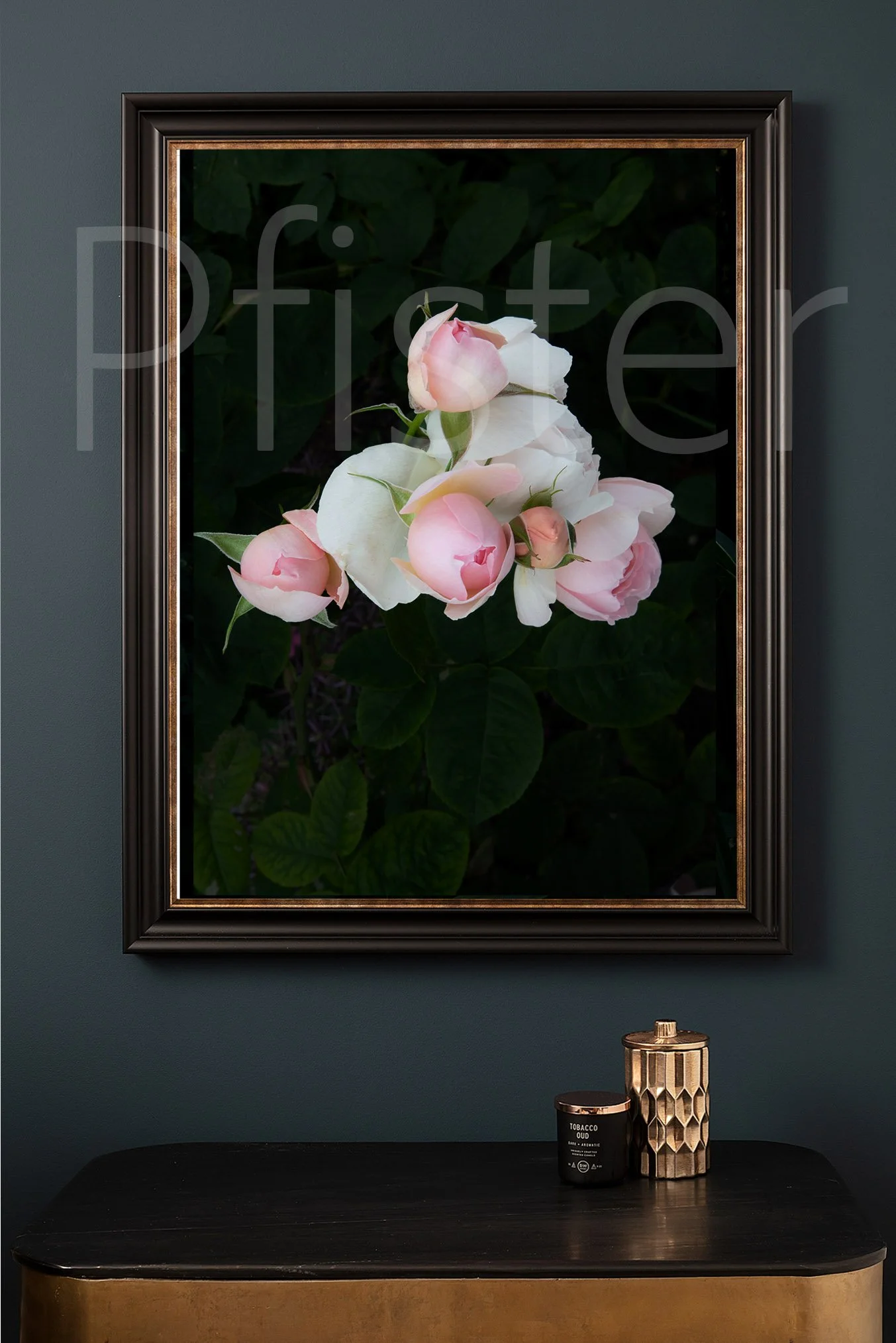 Framed photograph of pink and white peony flowers hanging on a dark green wall, with a black table holding a black candle labeled 'Tobacco Oud' and a tall gold and black geometric vase.