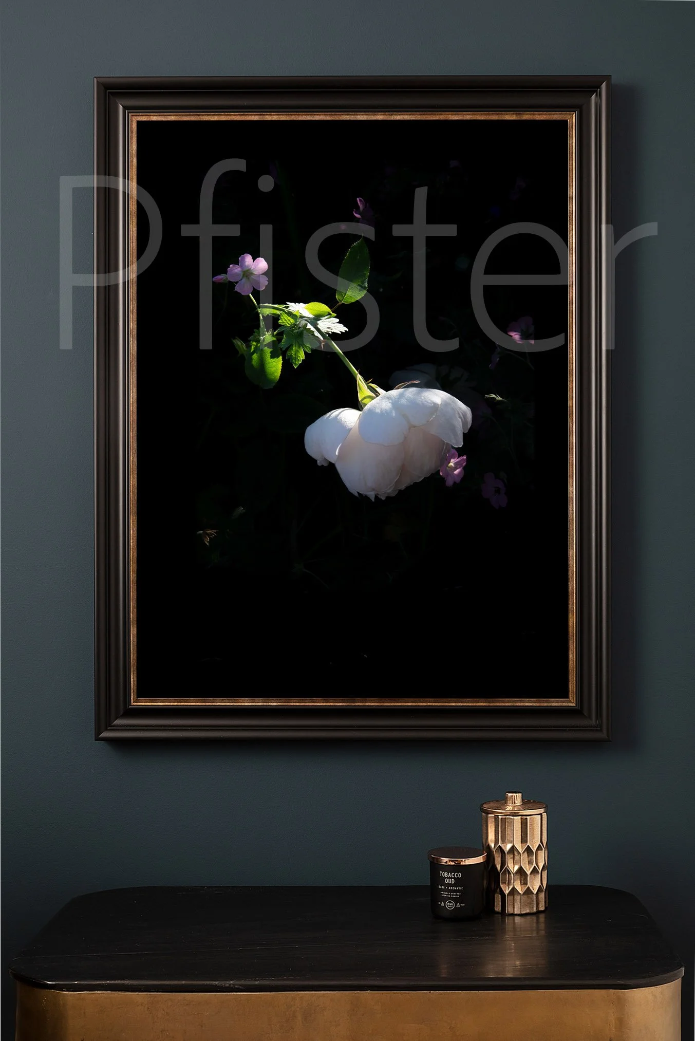 A framed floral artwork of a white flower with green leaves and purple accents hanging above a black and gold table, with a black candle and a decorative gold container on it.