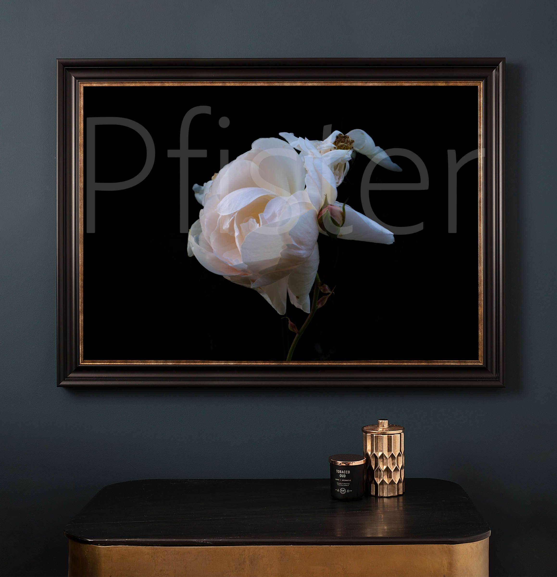 A framed photograph of a white peony flower displayed on a dark wall above a black table. On the table are a black candle and a gold textured container.