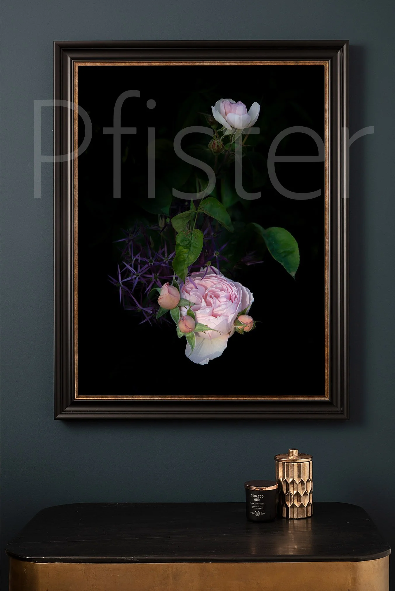 A framed photograph of pink and white roses on a dark background hangs on a dark green wall. A black table below holds a black candle labeled 'Tobacco Oud' and a geometric gold and black decorative container.