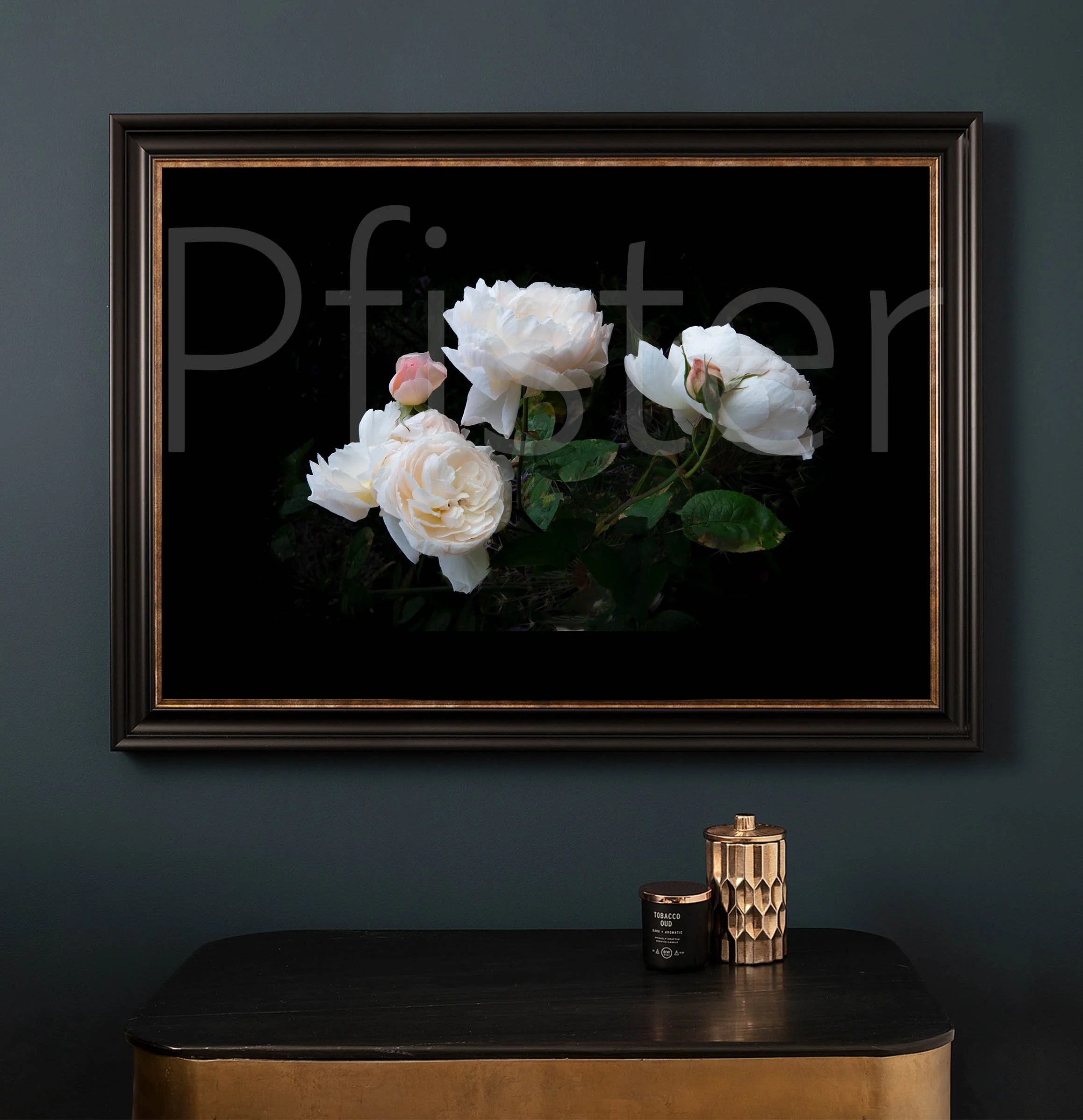 Black framed photograph of white roses on a dark background hanging on a dark green wall, with a black table below holding a candle and a decorative container.