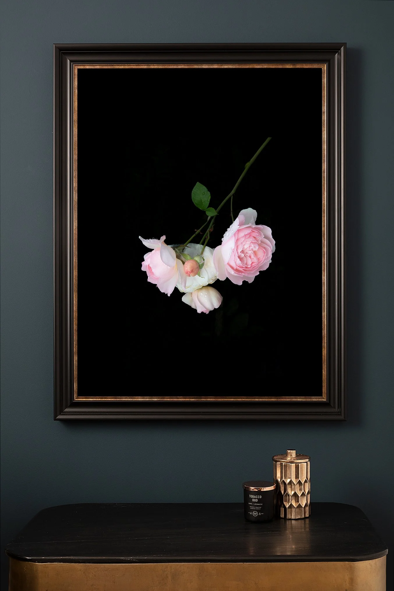 Decorative framed photo of pink and white flowers hanging on a dark green wall above a black and gold console table with a black candle and a decorative gold container.