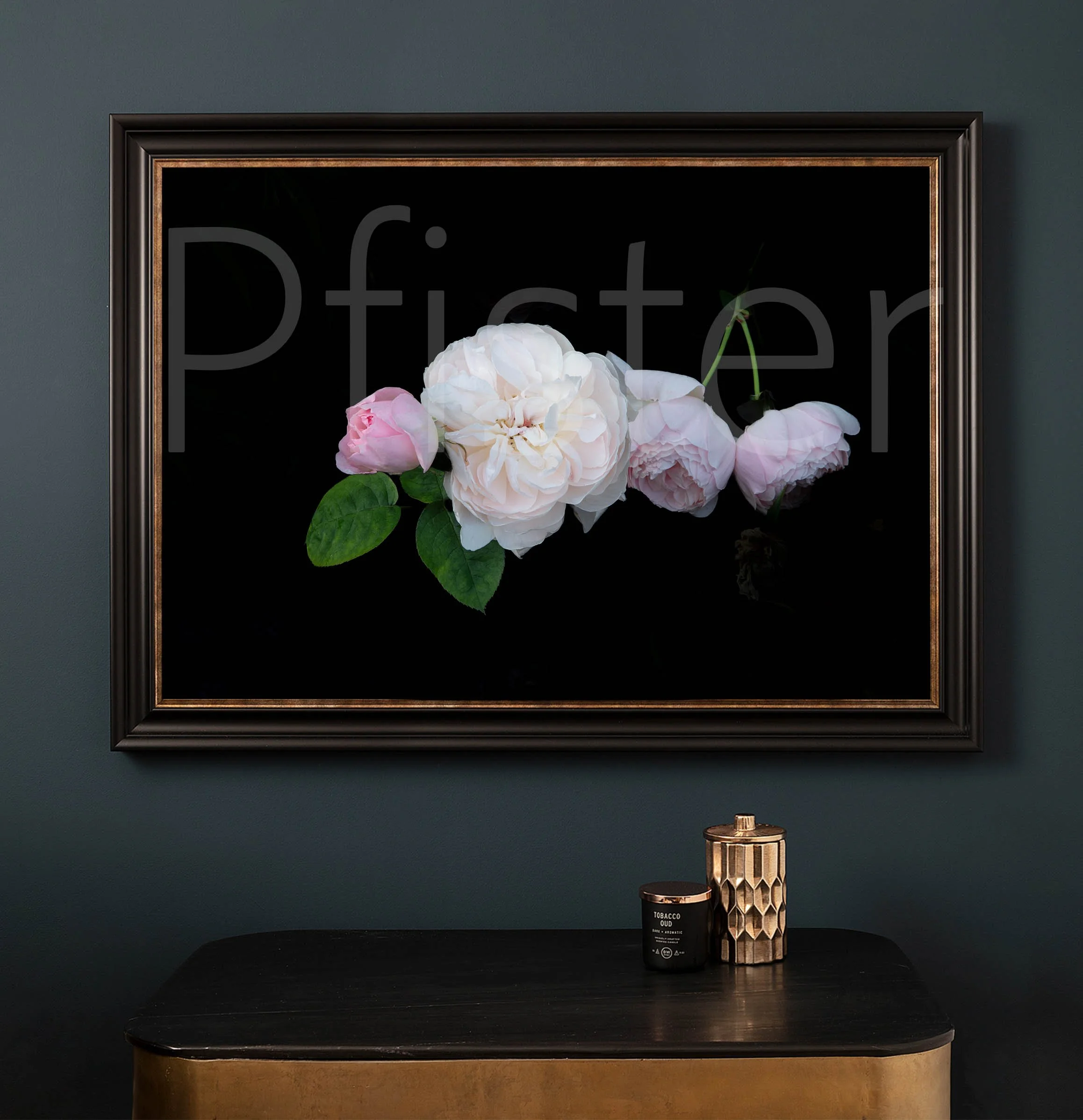 A black framed floral painting of pink and white roses on a dark wall, positioned above a black and gold table with a black candle and a decorative gold vase.