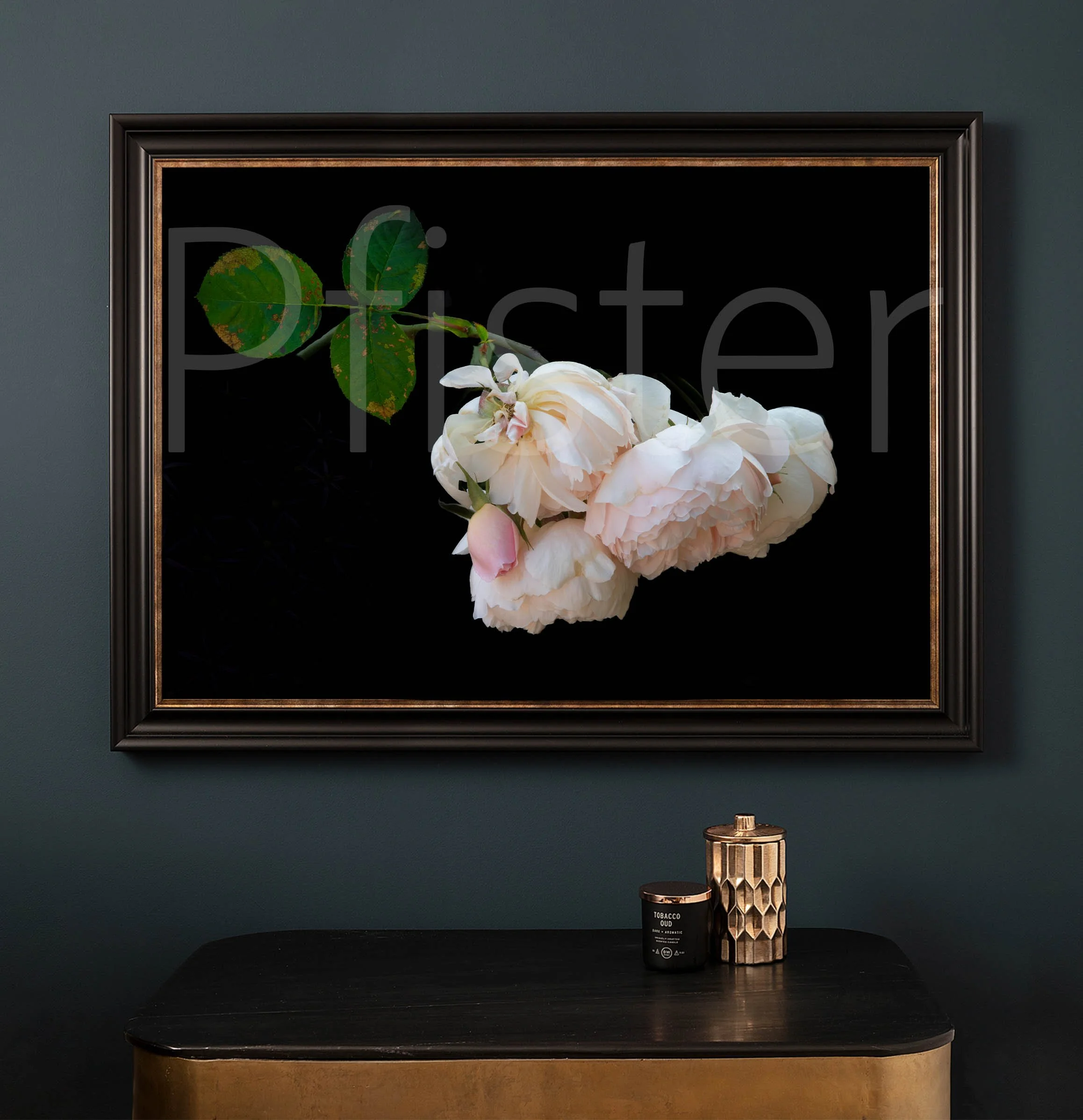 Framed floral artwork of white peonies hangs on a dark green wall above a black and gold table. On the table, there are a candle and a metallic decorative container.